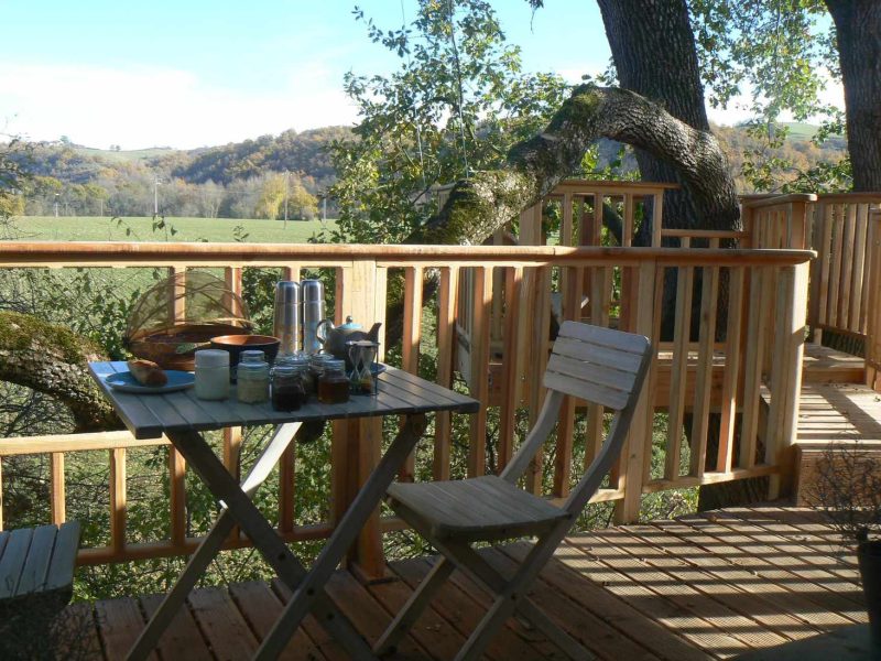 Cabane dans les arbres avec une terrasse en bois et une table pour le petit-déjeuner.