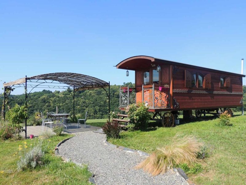 Roulotte en bois avec terrasse ombragée, entourée de verdure et de fleurs.