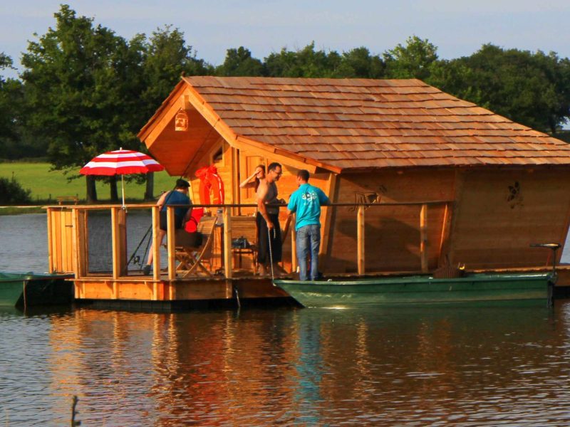 Cabane flottante en bois sur un lac, entourée de verdure et de canoës.