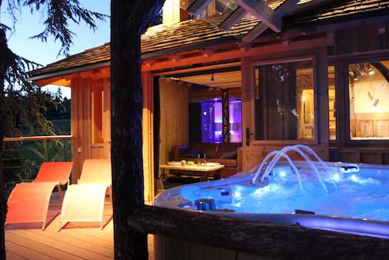 Chalet en bois avec jacuzzi illuminé et chaises longues sur la terrasse.