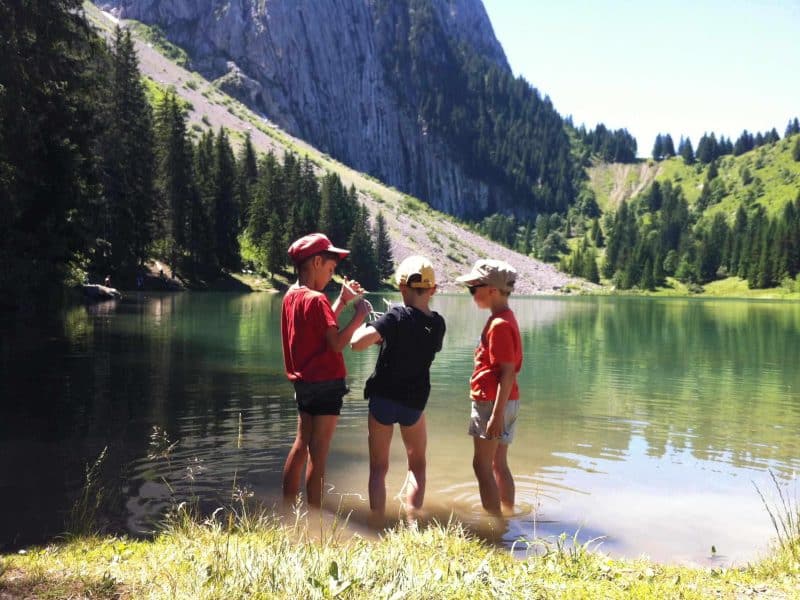 Trois enfants jouent au bord dun lac, entourés de montagnes verdoyantes.