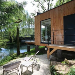 Cabane en bois sur pilotis au bord dun étang, entourée de verdure.