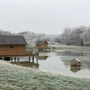 Cabane sur pilotis au bord dun lac gelé, entourée de paysages enneigés.