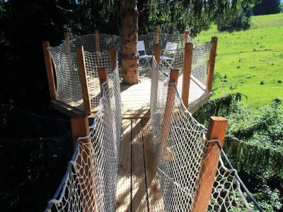 Cabane perchée dans un arbre, avec une passerelle en bois et vue sur la nature environnante.