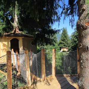 Cabane perchée dans les arbres, entourée de verdure et accessible par un pont en bois.
