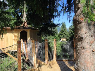 Cabane perchée dans les arbres, entourée de verdure et accessible par un pont en bois.