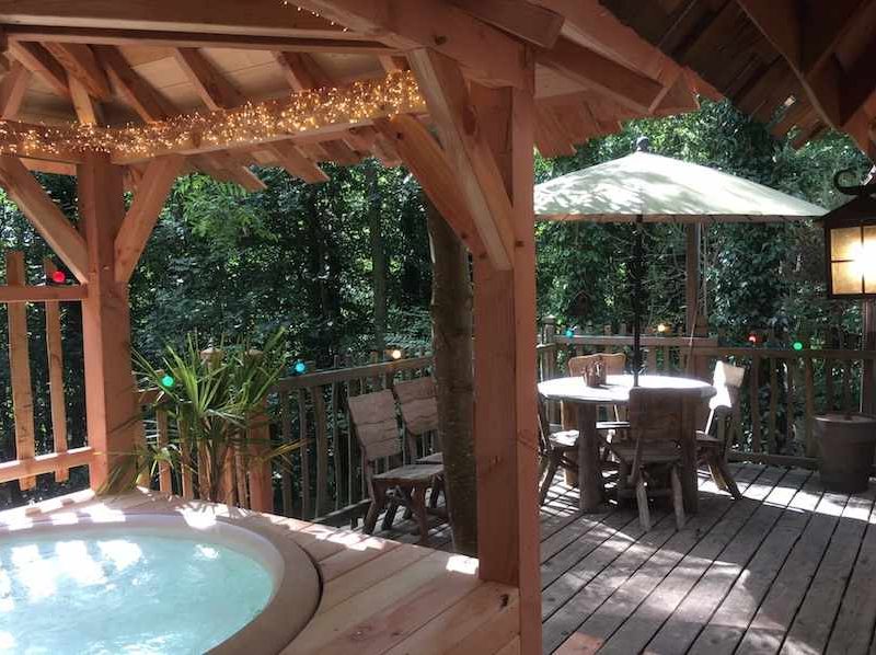 Cabane en bois avec jacuzzi, terrasse en bois entourée de verdure.