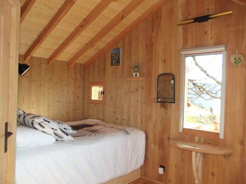 Cabane en bois avec lit douillet et vue sur la nature à travers une fenêtre.