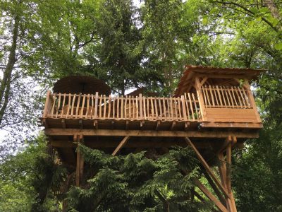 Cabane perchée dans les arbres, avec terrasse et parasol, entourée de verdure.