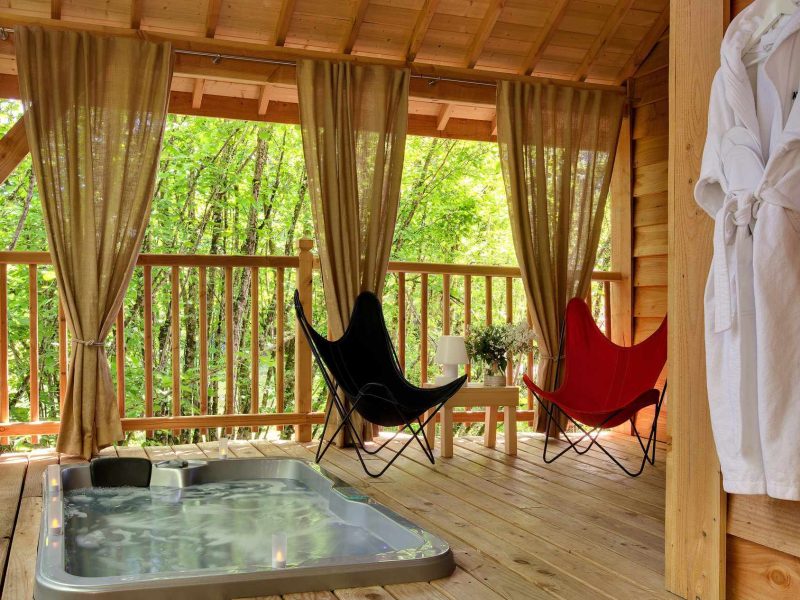 Cabane en bois avec jacuzzi, chaises confortables et vue sur la nature verdoyante.