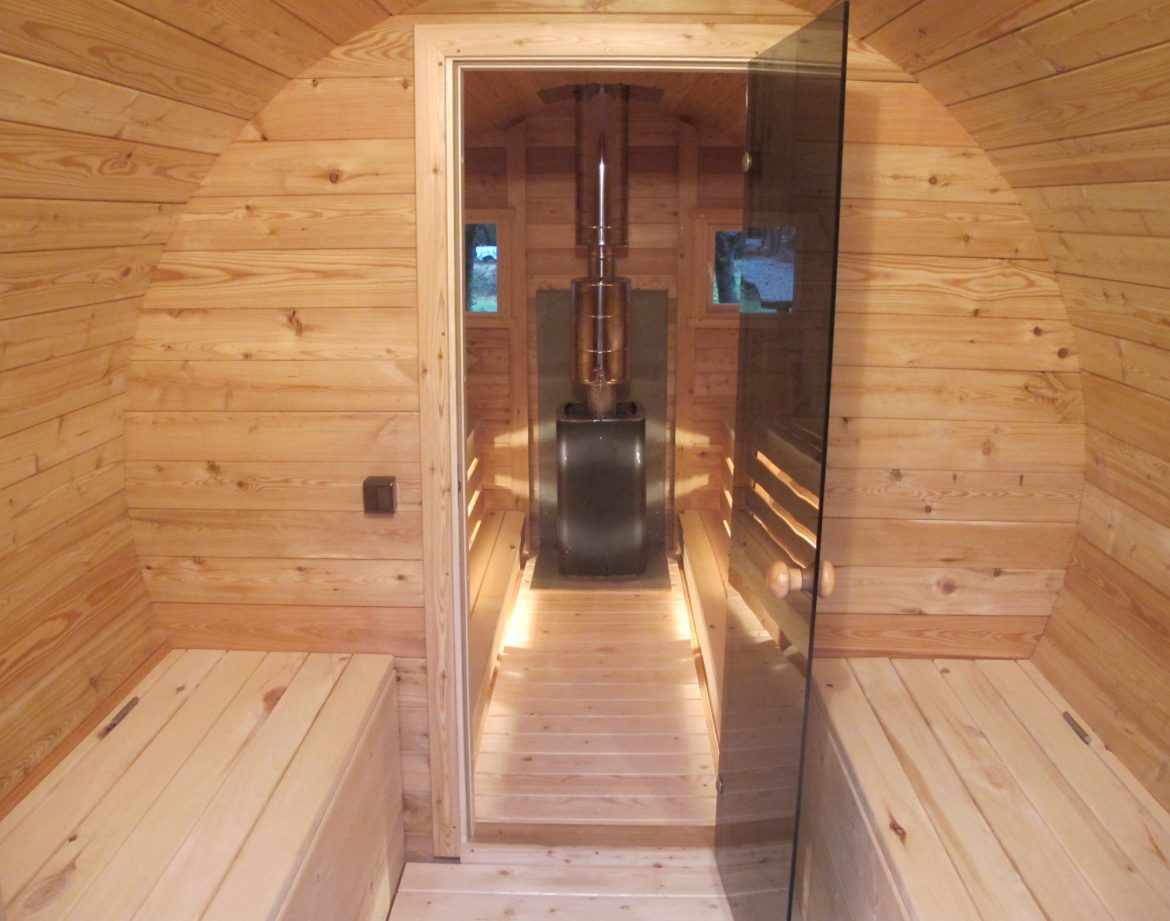 Hébergement insolite en forme de sauna en bois, avec un poêle central et un éclairage chaleureux.
