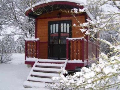 Roulotte en bois sous la neige, entourée darbres, offrant un cadre chaleureux et unique.