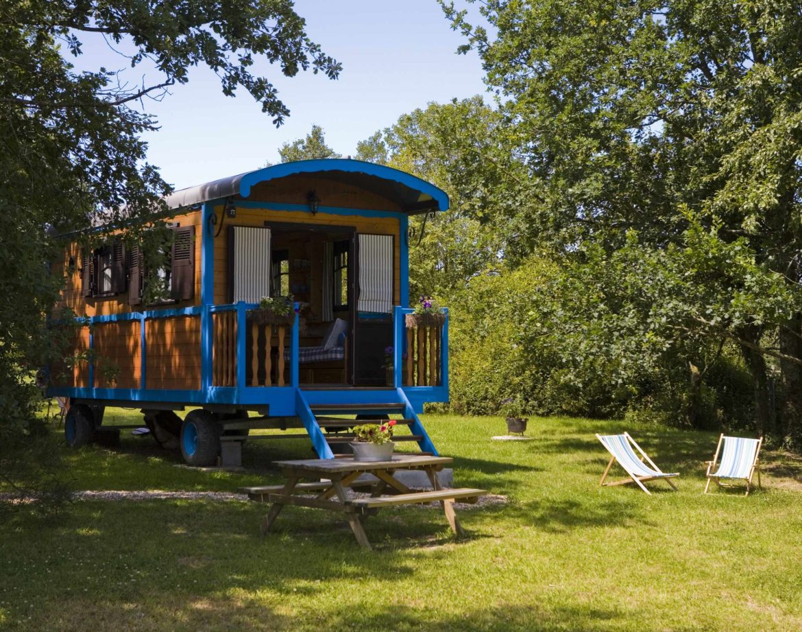 Charmante roulotte colorée entourée de verdure, avec chaises longues et table en bois.