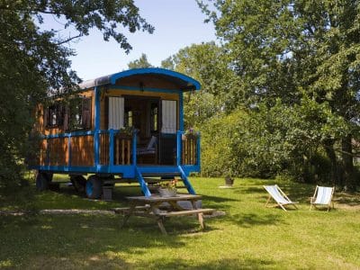 Charmante roulotte colorée entourée de verdure, avec chaises longues et table en bois.
