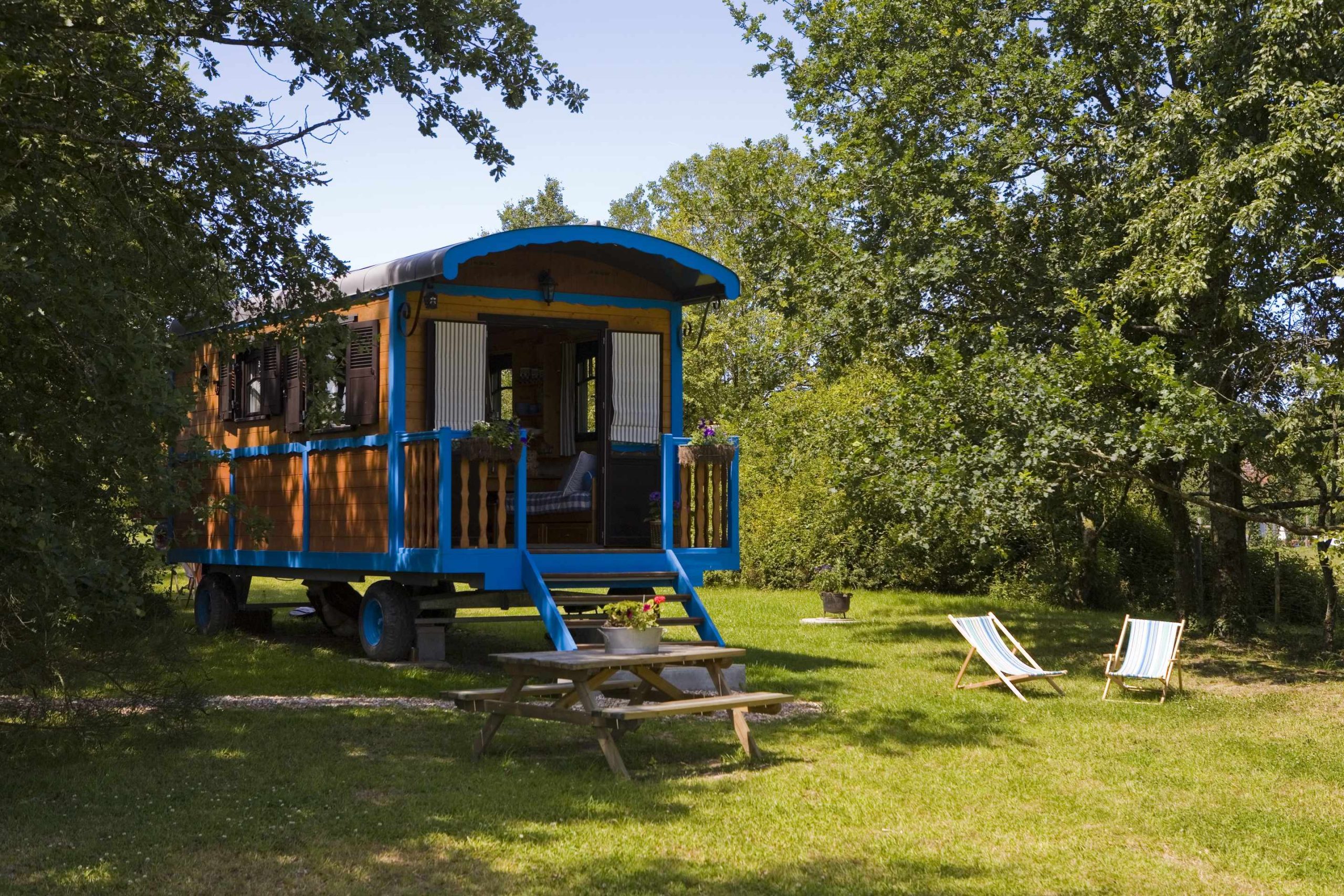 _MG_9512 Charmante roulotte colorée entourée de verdure, avec chaises longues et table en bois.