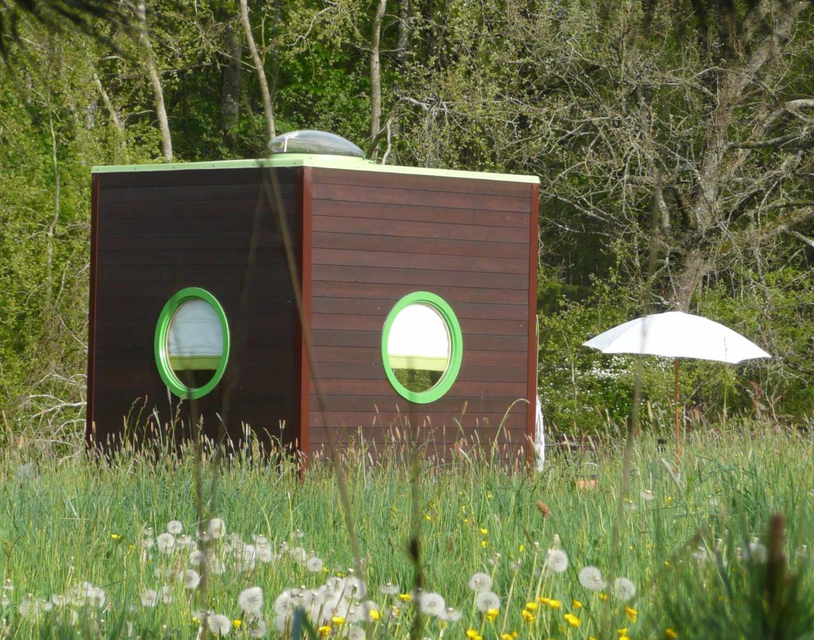 Hébergement insolite en forme de cube, avec fenêtres rondes, entouré de fleurs.