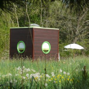 Hébergement insolite en forme de cube, avec fenêtres rondes, entouré de fleurs.