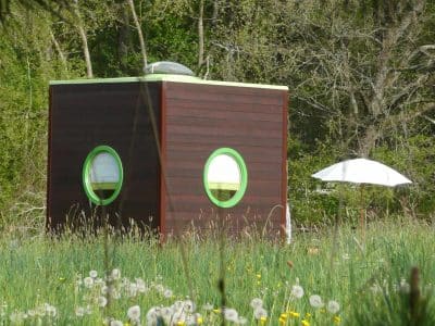 Hébergement insolite en forme de cube, avec fenêtres rondes, entouré de fleurs.