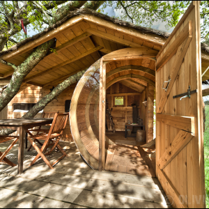 Cabane en bois perchée dans un arbre, avec une terrasse en bois et vue sur la nature.