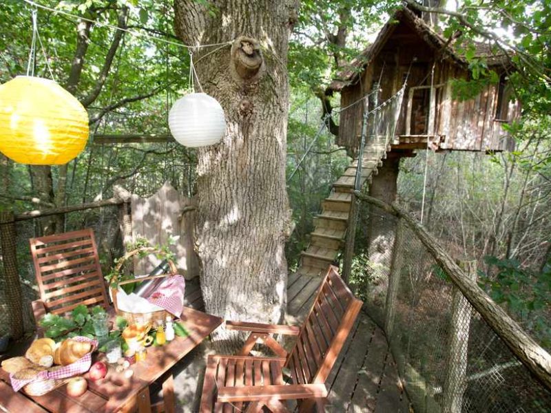 Cabane dans les arbres avec terrasse en bois, lanternes colorées et vue sur la forêt.