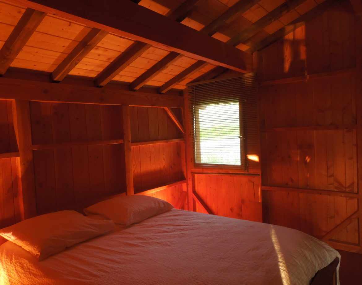 Chalet en bois chaleureux avec lit double et lumière douce filtrant par la fenêtre.