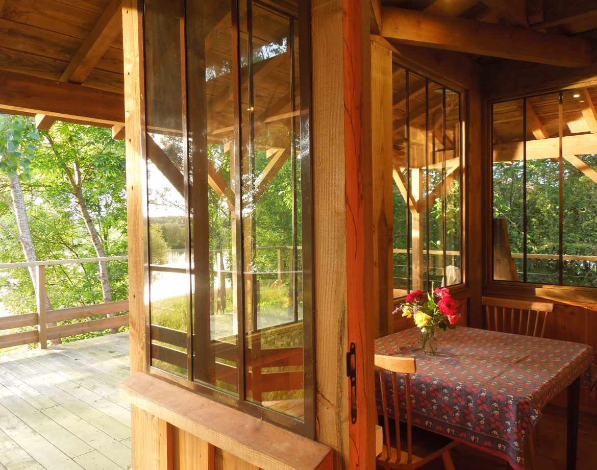 Cabane en bois avec grandes fenêtres, vue sur la nature et une table décorée de fleurs.