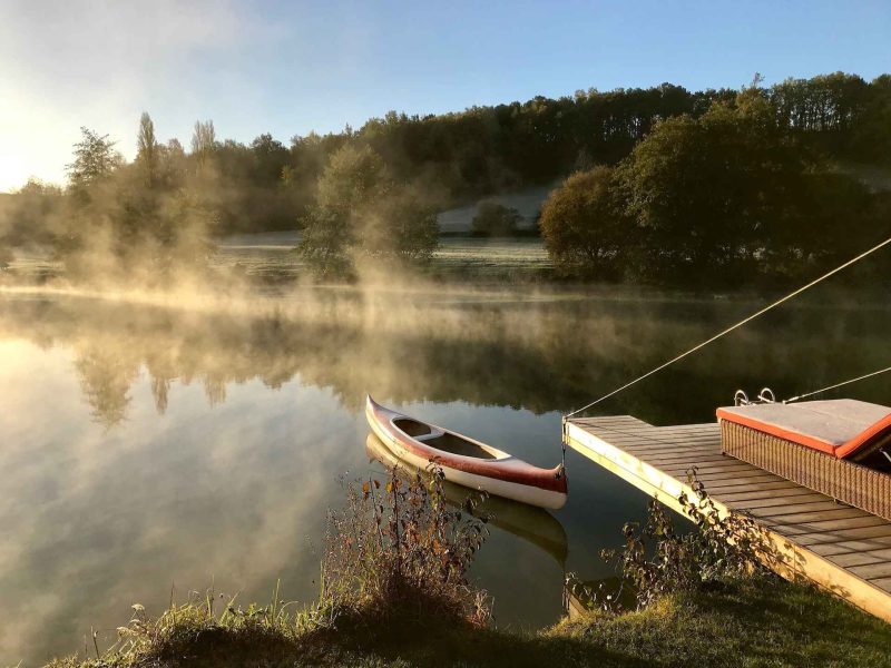 Cabane au bord de leau, avec un kayak amarré et une brume matinale.