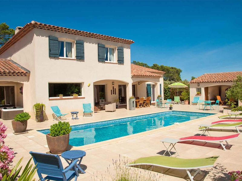 Villa avec piscine, entourée de fleurs et chaises longues sous un ciel bleu.