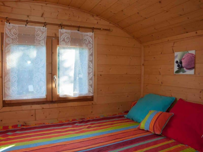 Chalet en bois avec lit coloré et fenêtres lumineuses, ambiance chaleureuse.