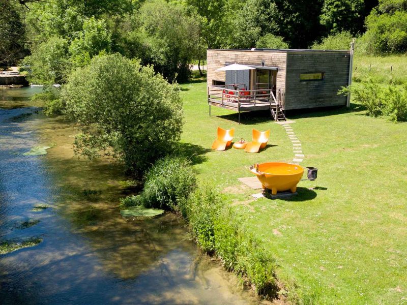Cabane moderne au bord de leau, avec chaises orange et verdure environnante.