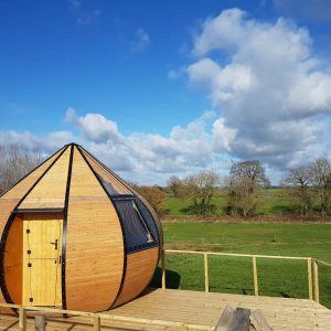 Cabane en bois en forme de goutte, entourée de verdure sous un ciel nuageux.
