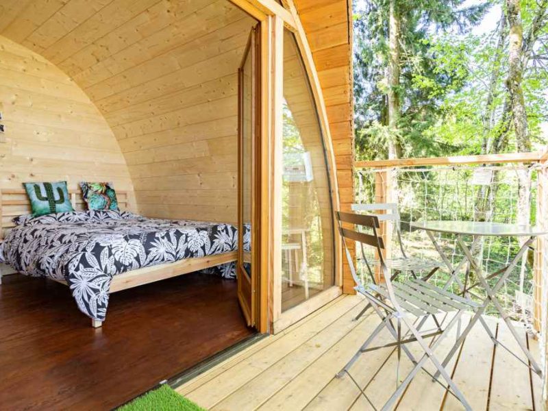Cabane en bois avec lit douillet et terrasse en bois entourée de verdure.
