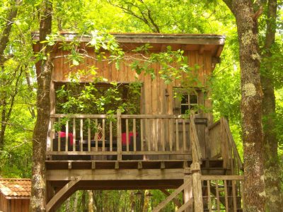 Cabane perchée en bois, entourée darbres verdoyants, avec balcon et fauteuils.