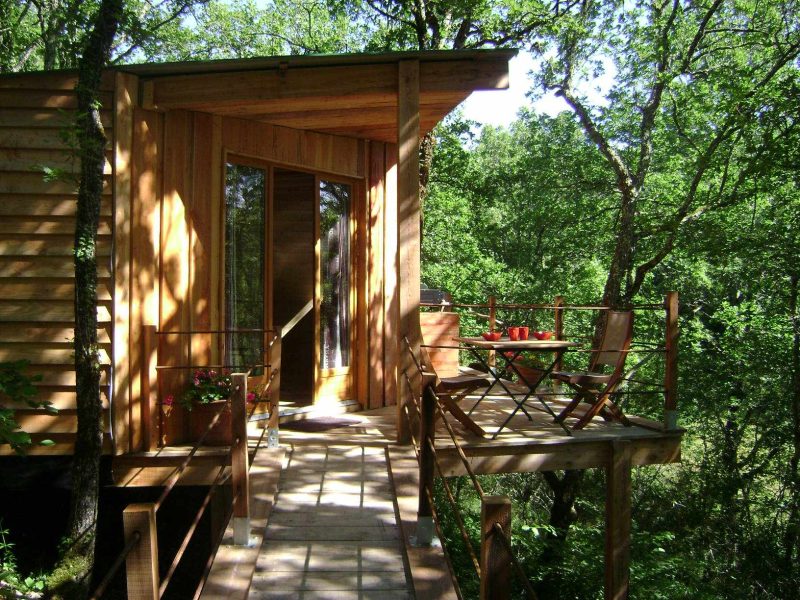 Cabane en bois perchée, entourée darbres, avec terrasse et mobilier extérieur.