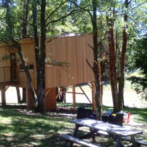 Cabane perchée dans les arbres, entourée de verdure, avec une terrasse en bois.