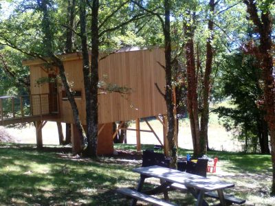 Cabane perchée dans les arbres, entourée de verdure, avec une terrasse en bois.