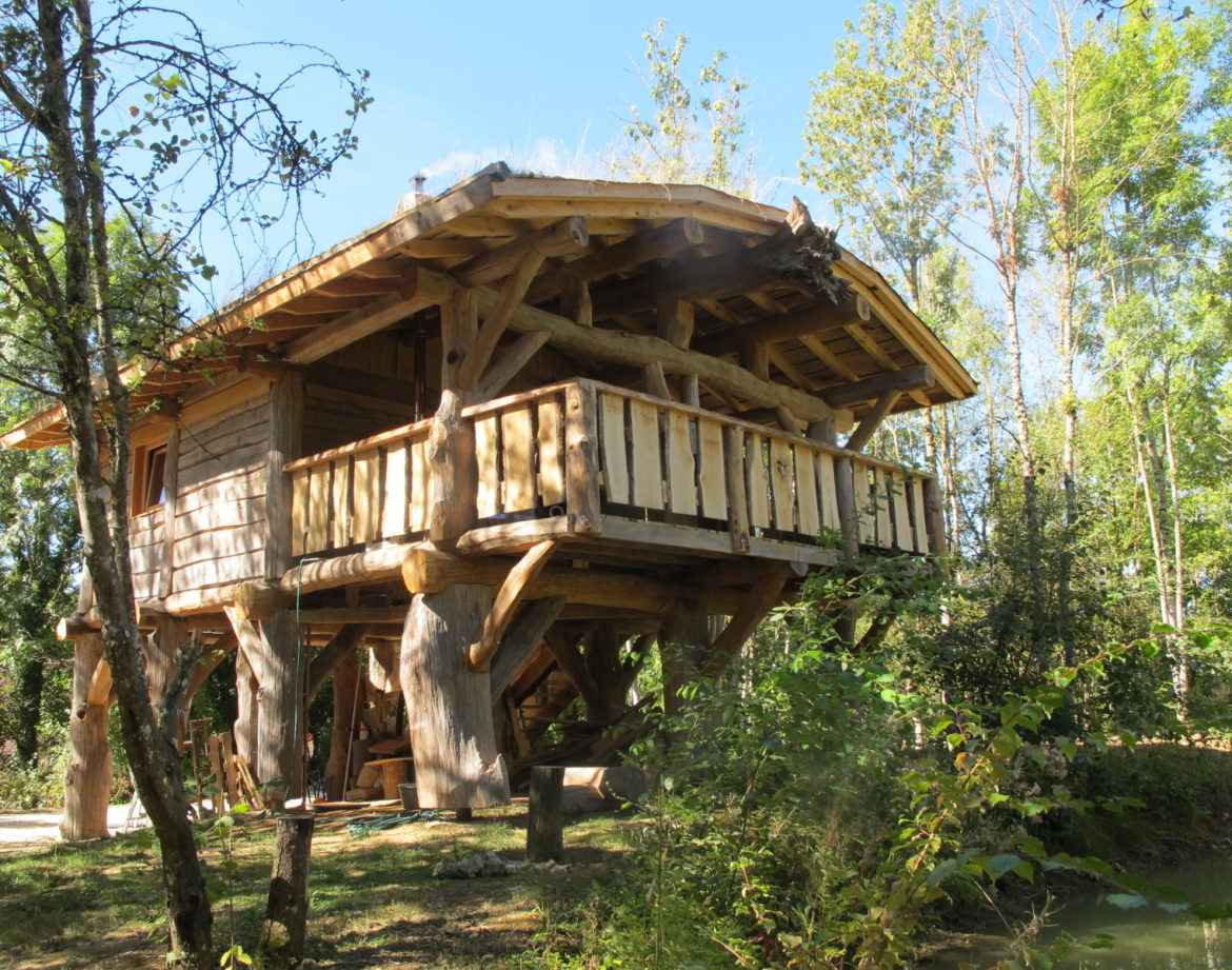 Cabane perchée en bois dans les arbres, entourée de verdure et dun ruisseau.