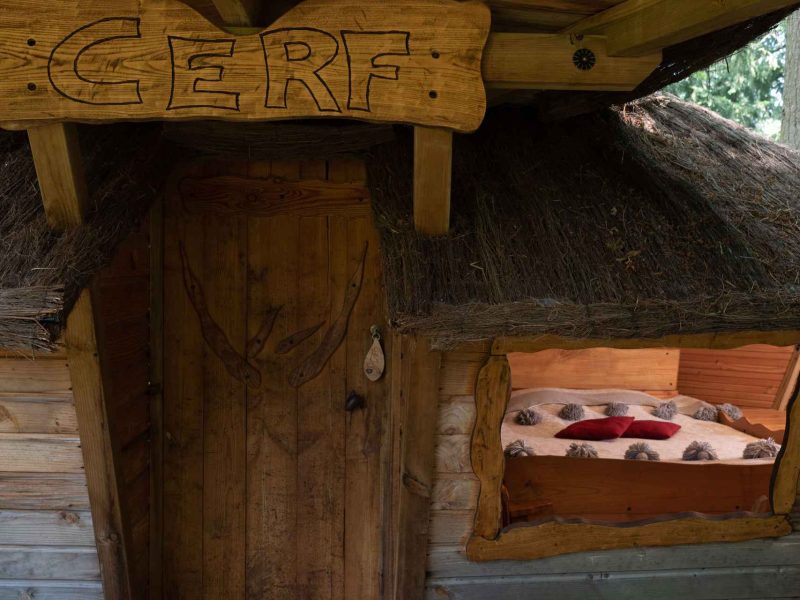Cabane en bois avec toit de chaume, lit douillet visible à travers la fenêtre.