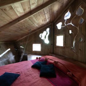 Cabane en bois avec lit douillet et fenêtres artistiques, située en Auvergne-Rhône-Alpes.