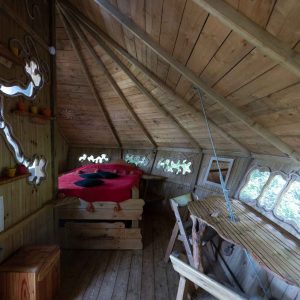 Cabane en bois avec un lit rouge et de grandes fenêtres, offrant une vue sur la nature.