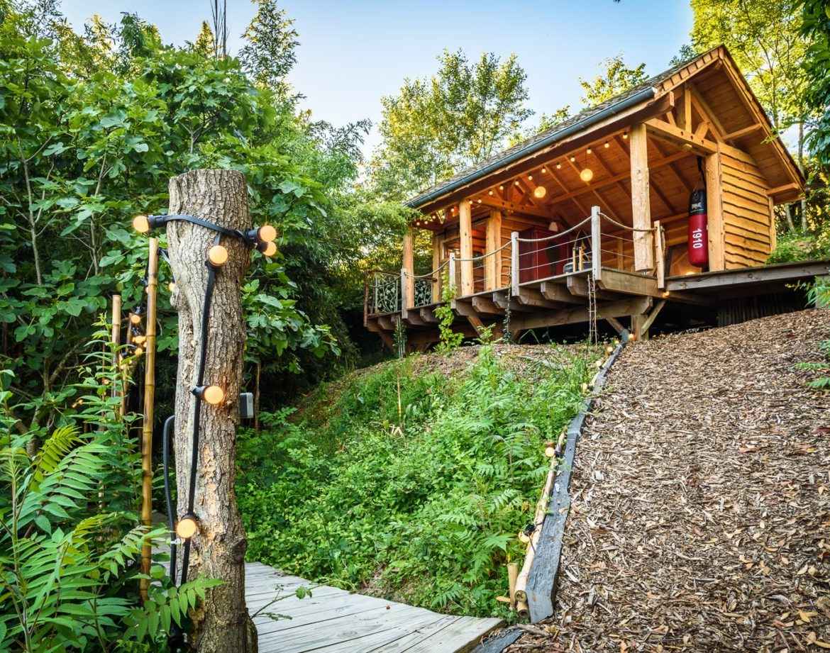 Cabane en bois perchée, entourée de verdure, avec guirlandes lumineuses.