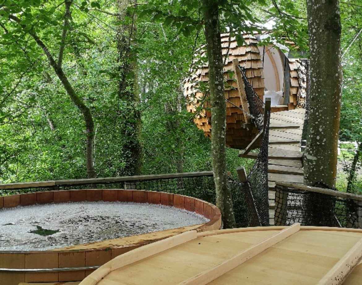 Cabane perchée dans les arbres avec un spa en bois, entourée de verdure luxuriante.