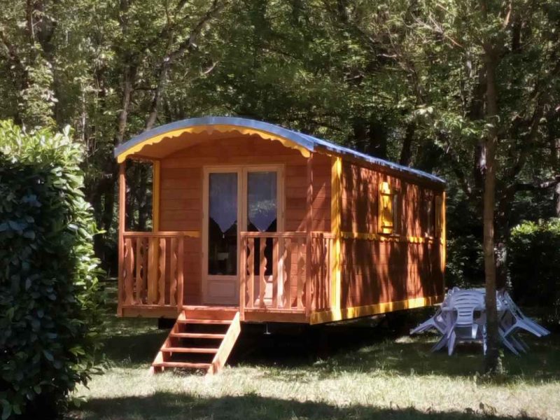 Cabane en bois colorée, entourée darbres, avec terrasse ensoleillée.