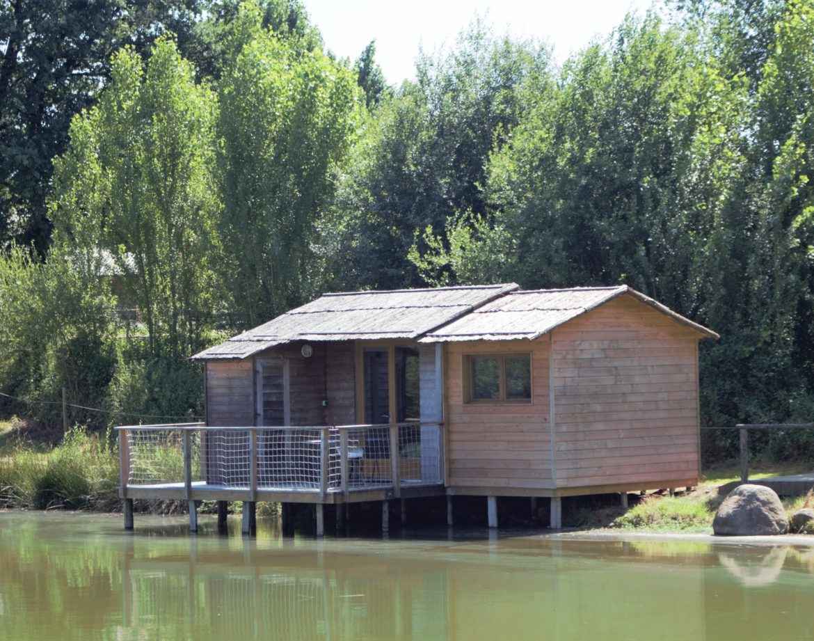 Cabane en bois sur pilotis au bord dun étang, entourée de verdure luxuriante.