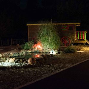 Roulotte colorée illuminée, entourée de verdure et dun petit bassin.