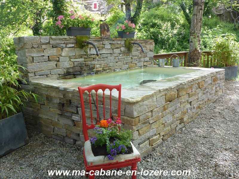 Cabane en Lozère avec un bassin en pierre et des fleurs colorées.