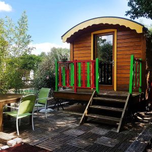 Roulotte colorée en bois, entourée de verdure, avec terrasse et mobilier extérieur.