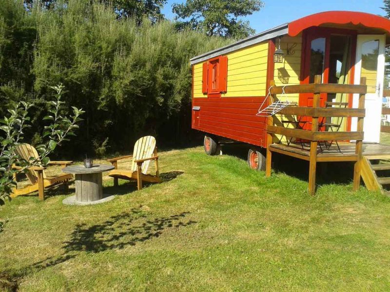 Charmante roulotte colorée avec terrasse et chaises en bois dans un cadre verdoyant.