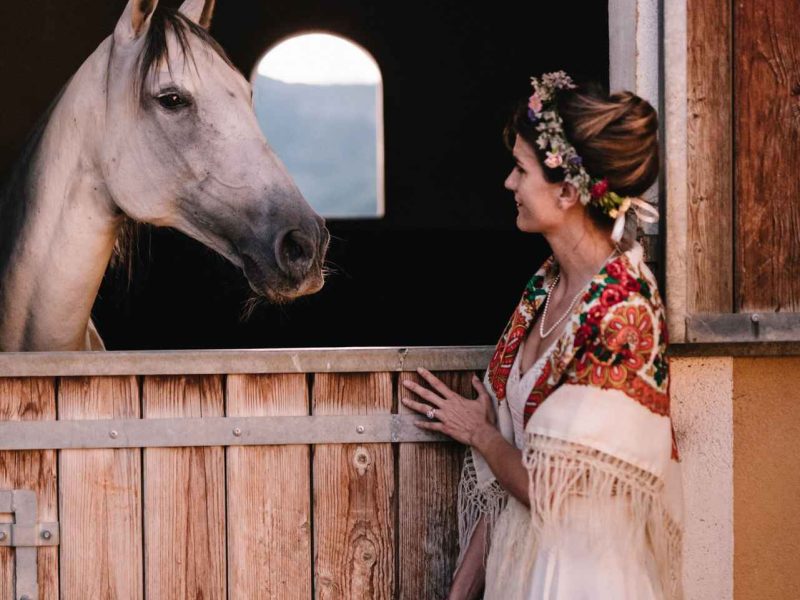 Hébergement équestre : une mariée souriante face à un cheval dans une écurie charmante.