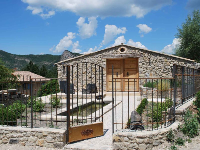 Gîte en pierre avec jardin, entouré de montagnes et ciel bleu.